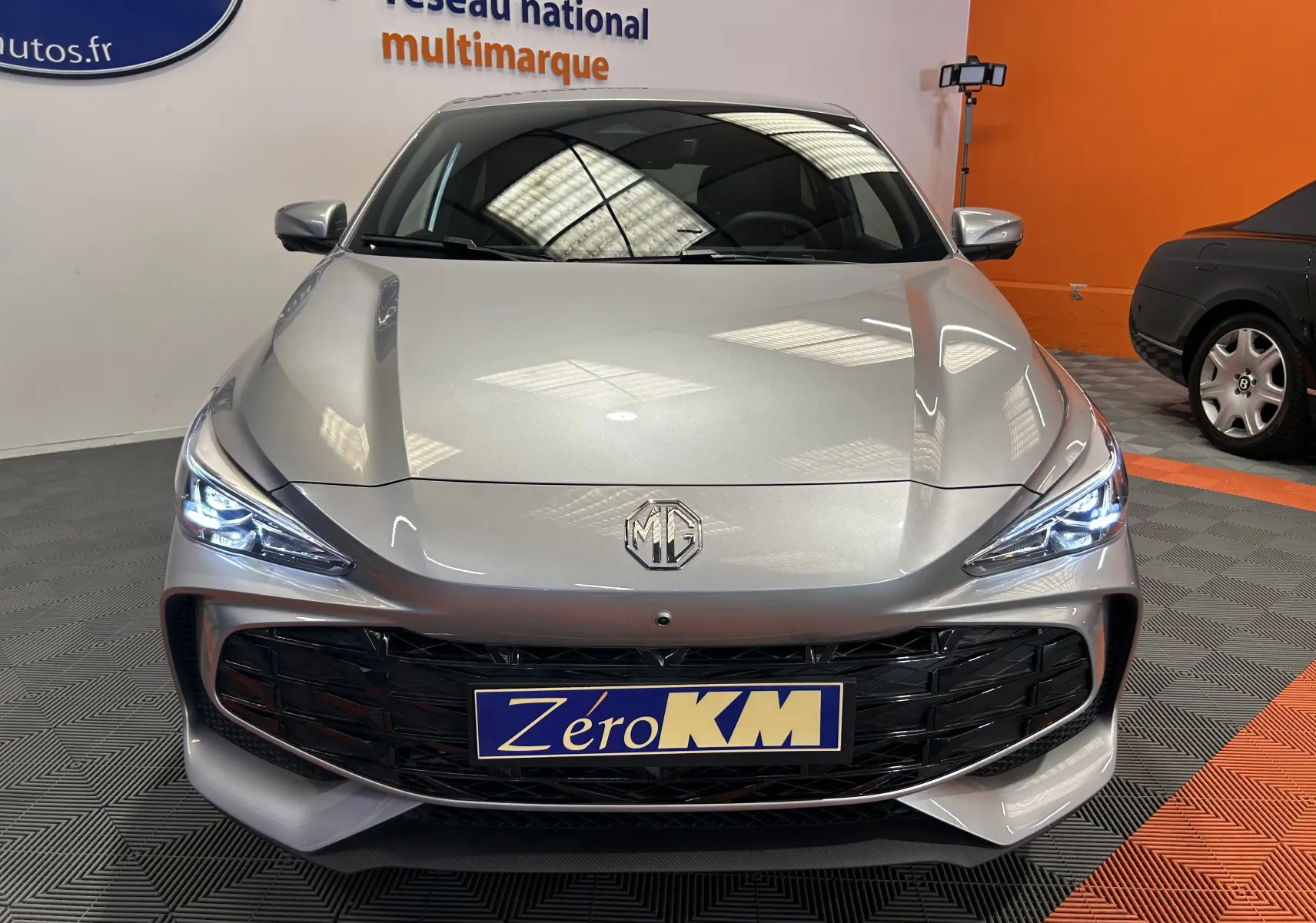 Vue avant d'une MG3 Cosmic Silver 2026 avec phares LED allumés et calandre noire brillante en intérieur showroom.