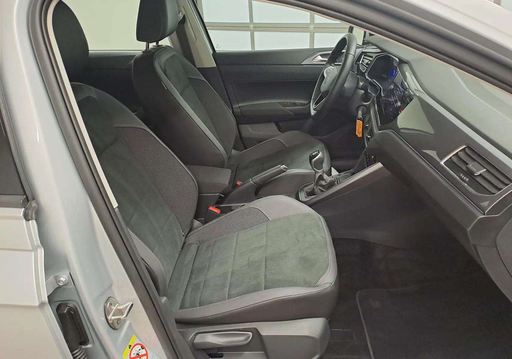 Vue intérieure côté conducteur de la Volkswagen Polo 2025 reflet d'argent, sièges noirs et levier de vitesses manuel visible.