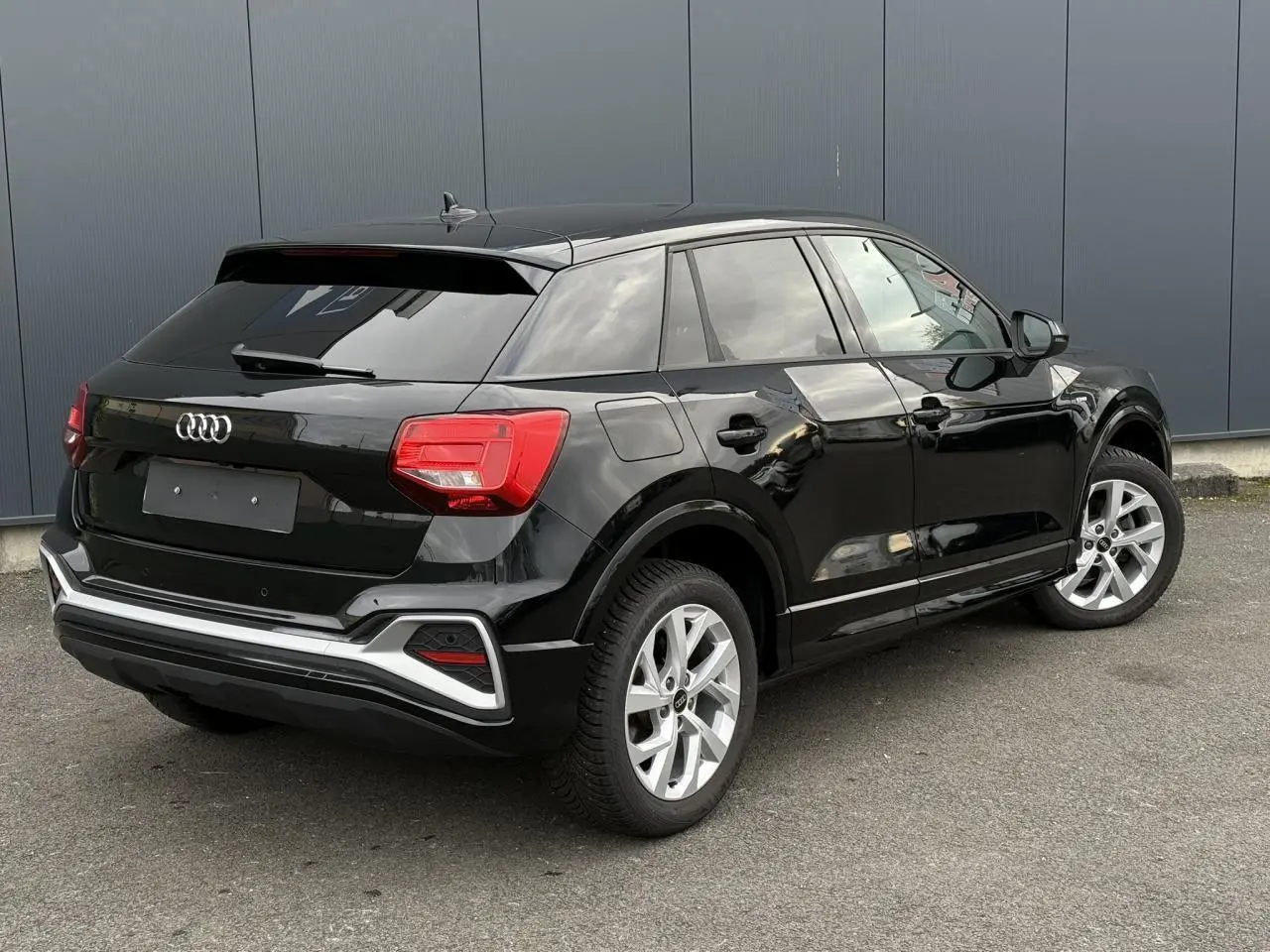 Audi Q2 noir métallisé vue 3/4 arrière droit, hayon électrique et jantes alliage visibles sur parking extérieur.