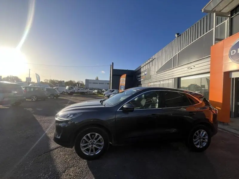 Ford Kuga 2.0 EcoBlue gris métallisé vu de profil côté gauche, stationné devant un bâtiment commercial.