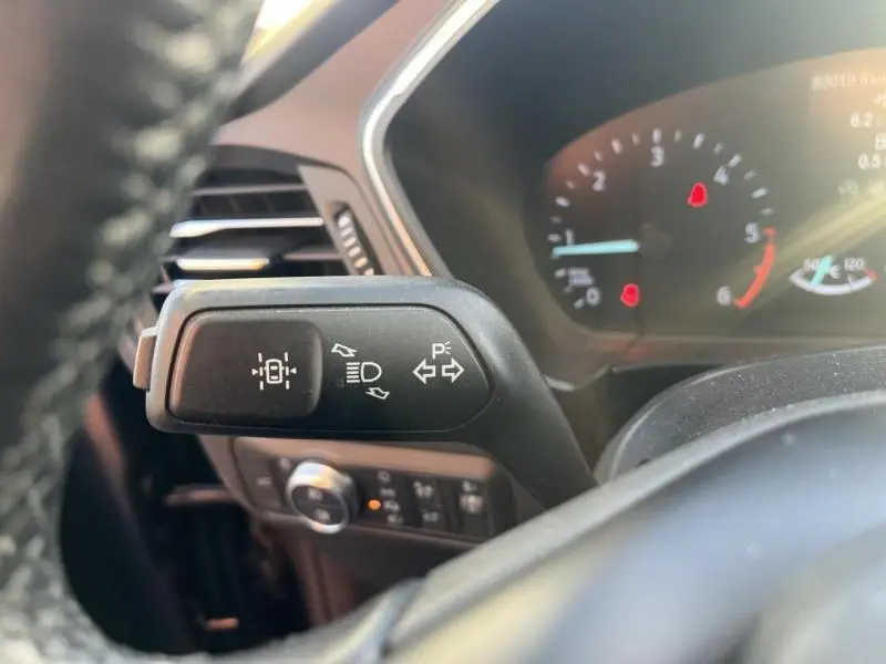 Gros plan sur la manette de clignotants à gauche du volant dans l'habitacle du Ford Kuga gris, avec tableau de bord numérique flou en arrière-plan.