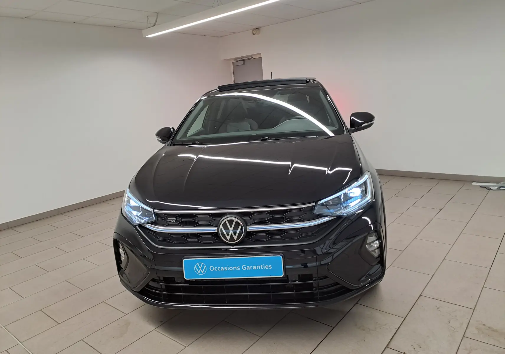 Vue avant d'un Volkswagen Taigo noir intense nacré avec calandre R-Line et phares LED allumés en intérieur showroom.