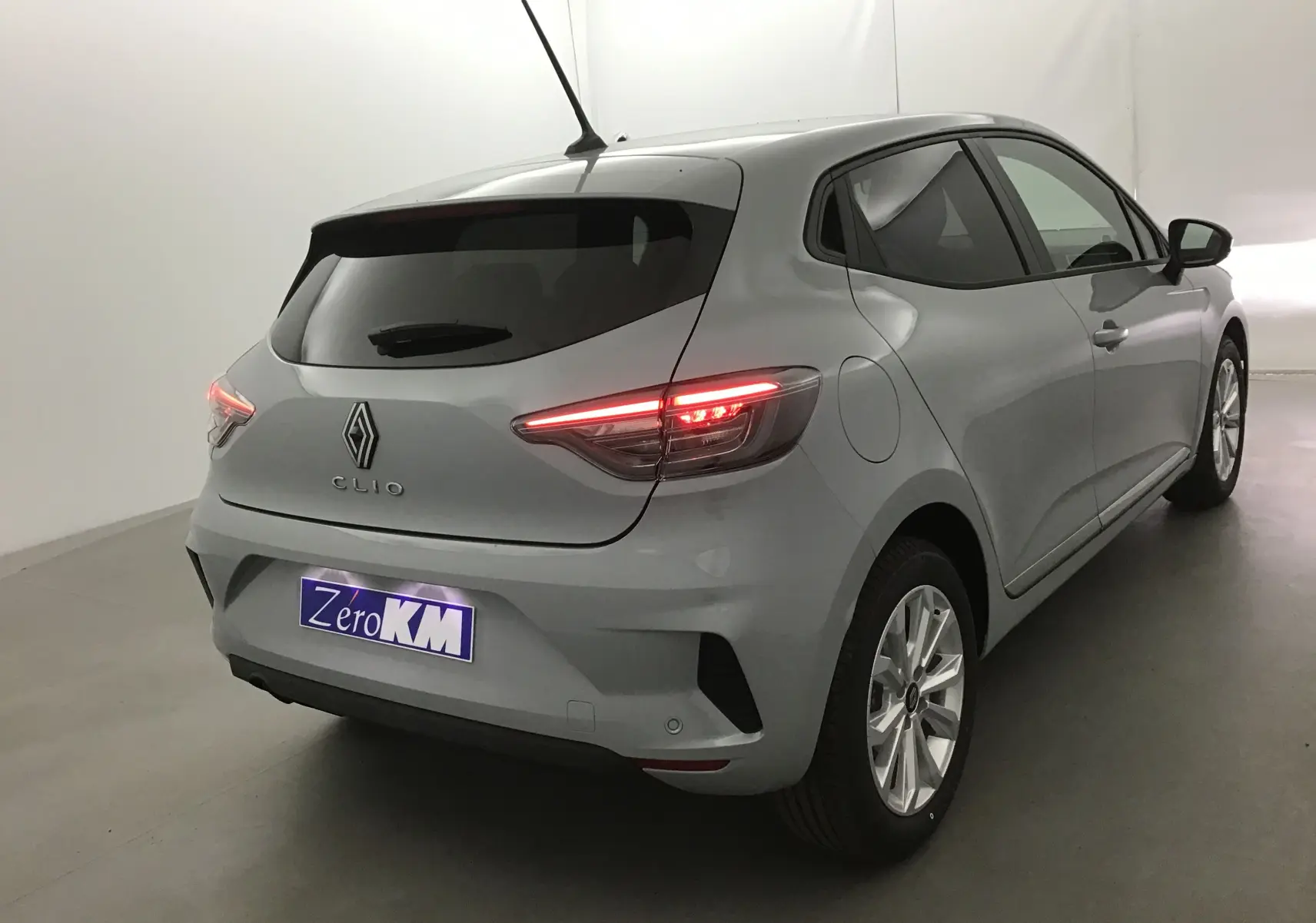 Vue 3/4 arrière droite d'une Renault Clio V gris Rafale 2025 avec feux arrière allumés et protections chromées latérales.
