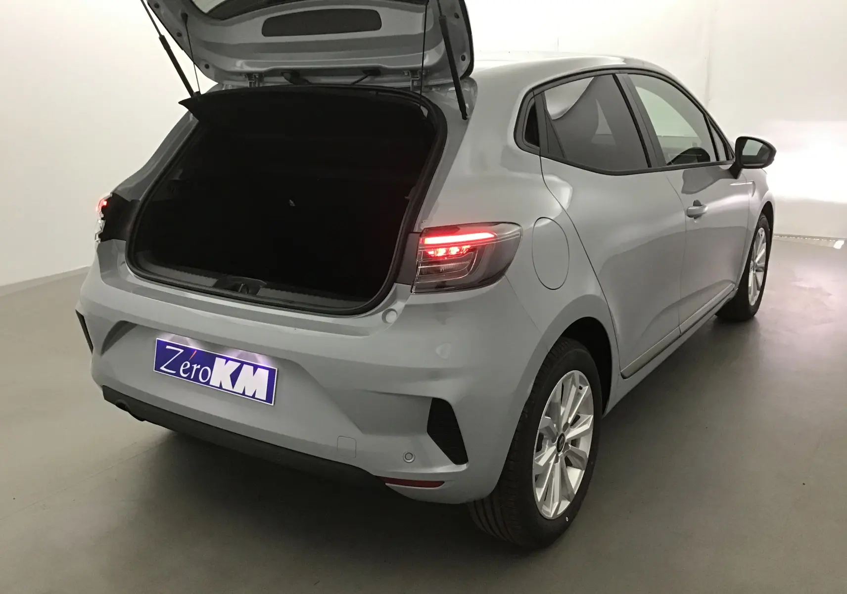 Vue 3/4 arrière droite d'une Renault Clio V gris Rafale avec coffre ouvert et feux arrière allumés dans un studio.