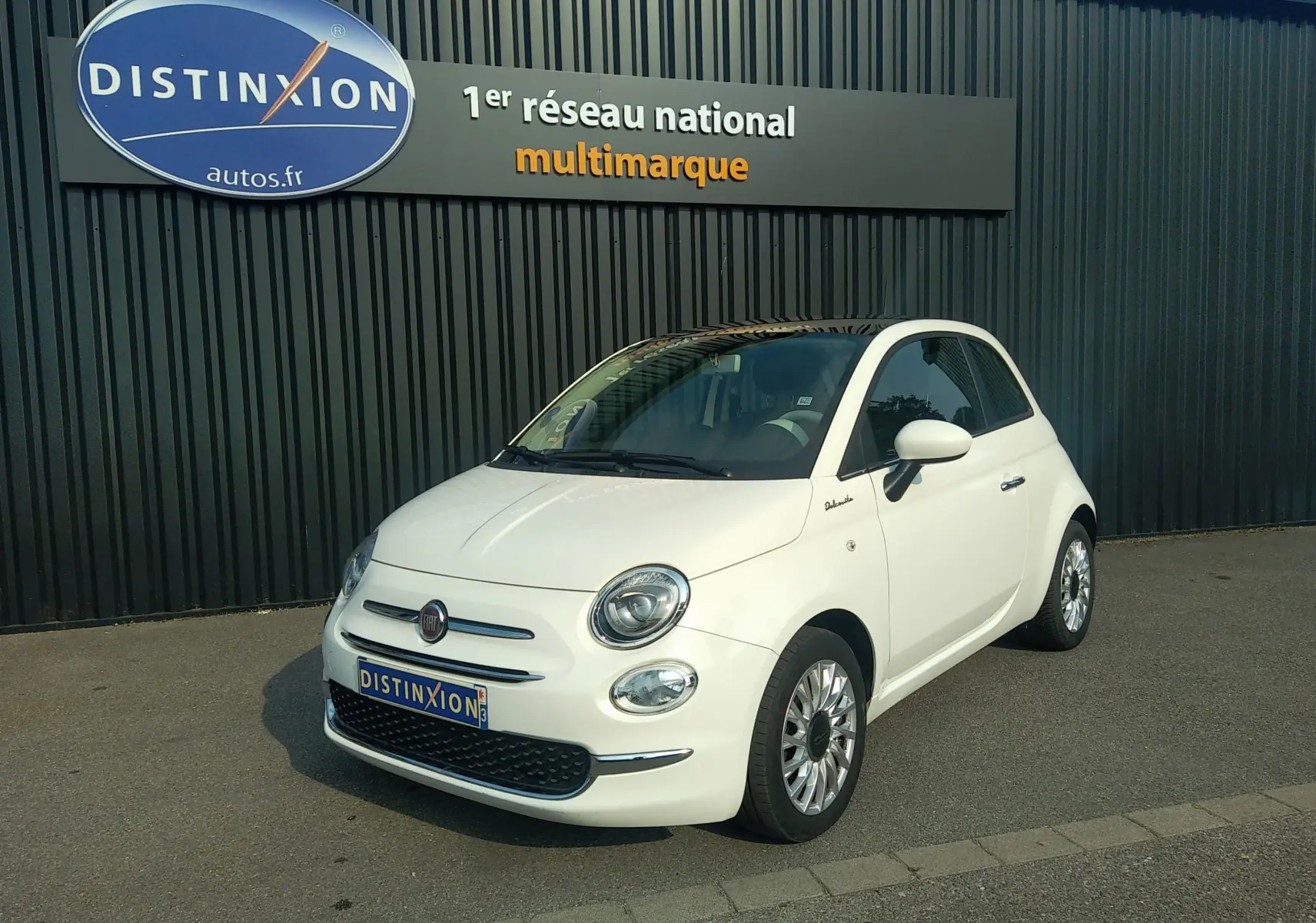 Fiat 500 blanche vue en 3/4 avant droit, avec toit noir et jantes alliage, stationnée devant un mur sombre.