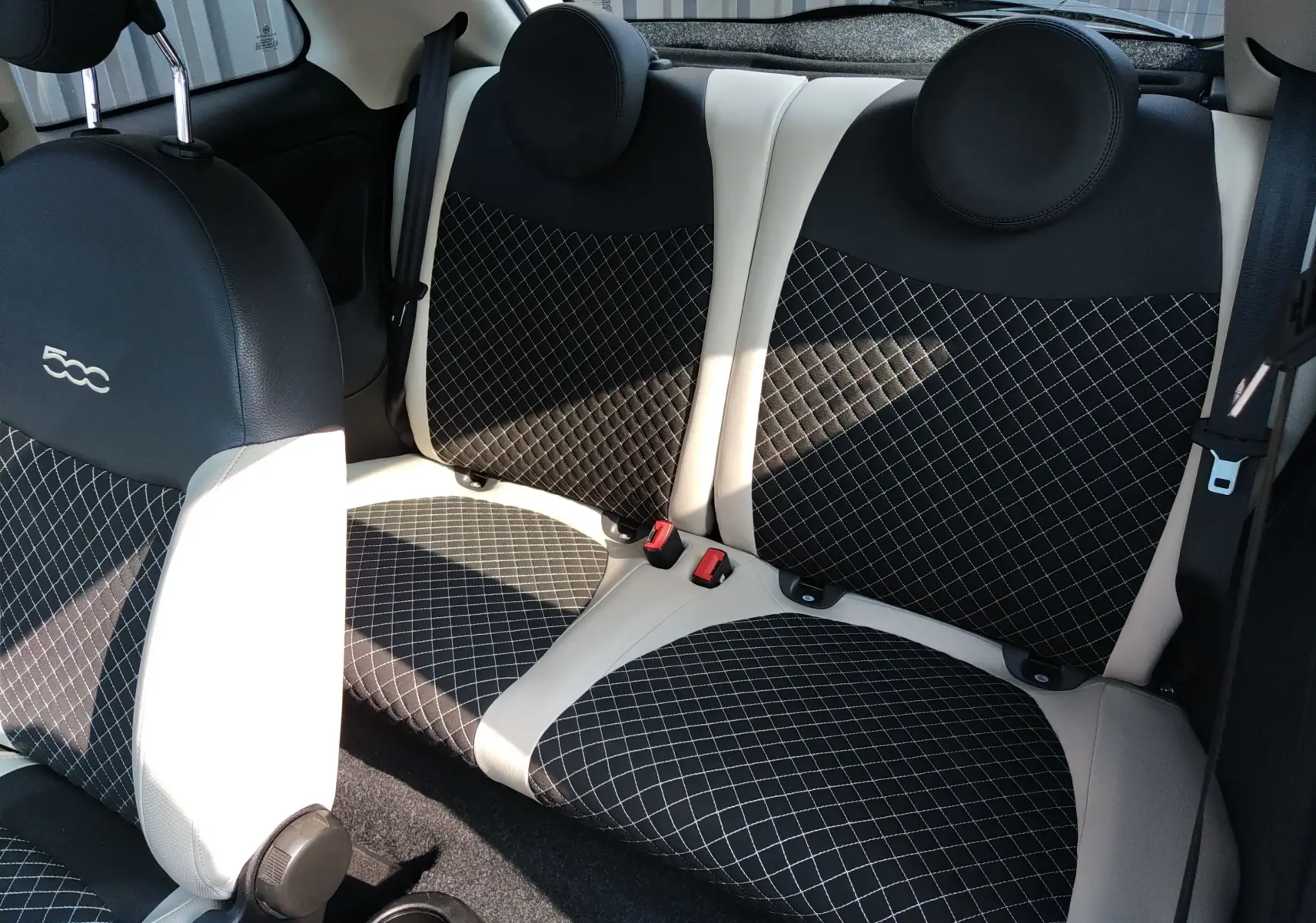 Vue intérieure arrière de la Fiat 500 blanche, sièges en tissu noir à motif quadrillé avec surpiqûres blanches et appuie-têtes ronds.