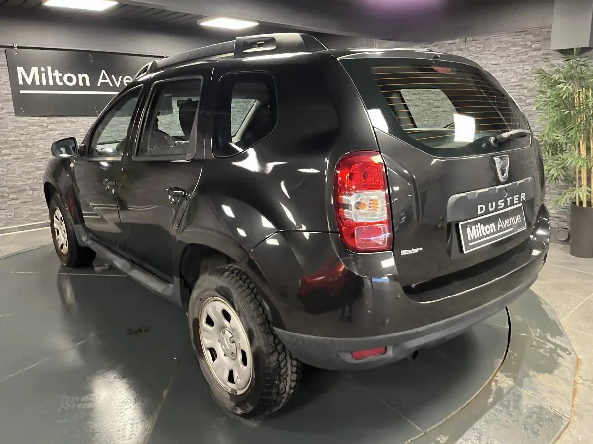 Vue 3/4 arrière gauche d'un Dacia Duster noir 2016 avec barres de toit et jantes acier en intérieur showroom.