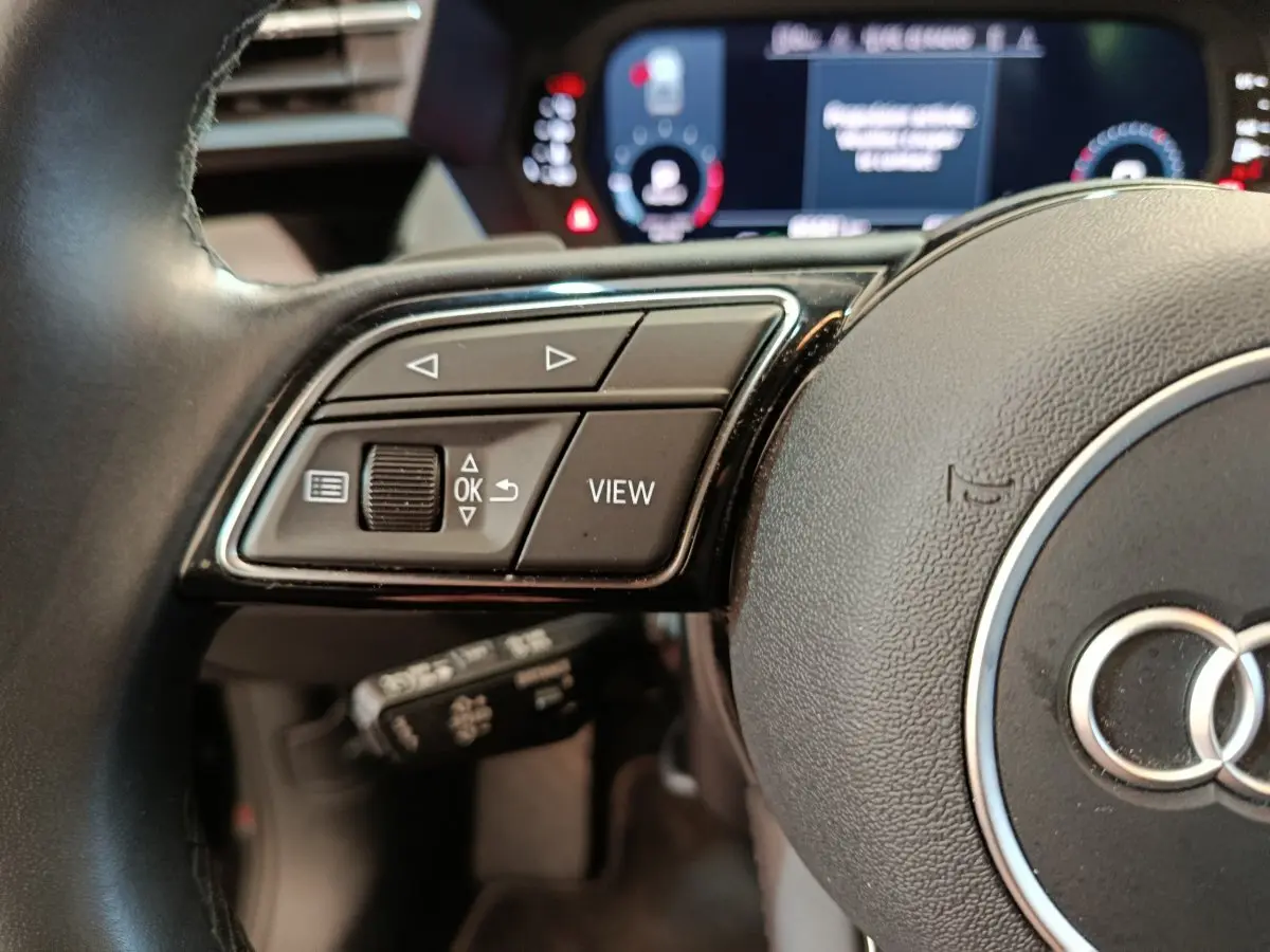 Gros plan sur les commandes gauche du volant cuir d'une Audi A3 noire, avec tableau de bord numérique flou en arrière-plan