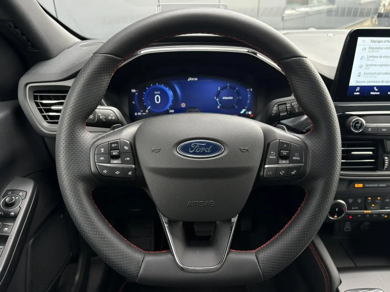Vue centrée sur le volant perforé noir avec surpiqûres rouges du Ford Kuga 2023, tableau de bord numérique et console tactile visibles.