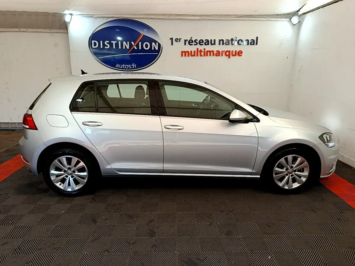 Profil droit d'une Volkswagen Golf gris clair 2019 garée en intérieur avec logo Distinxion en arrière-plan.