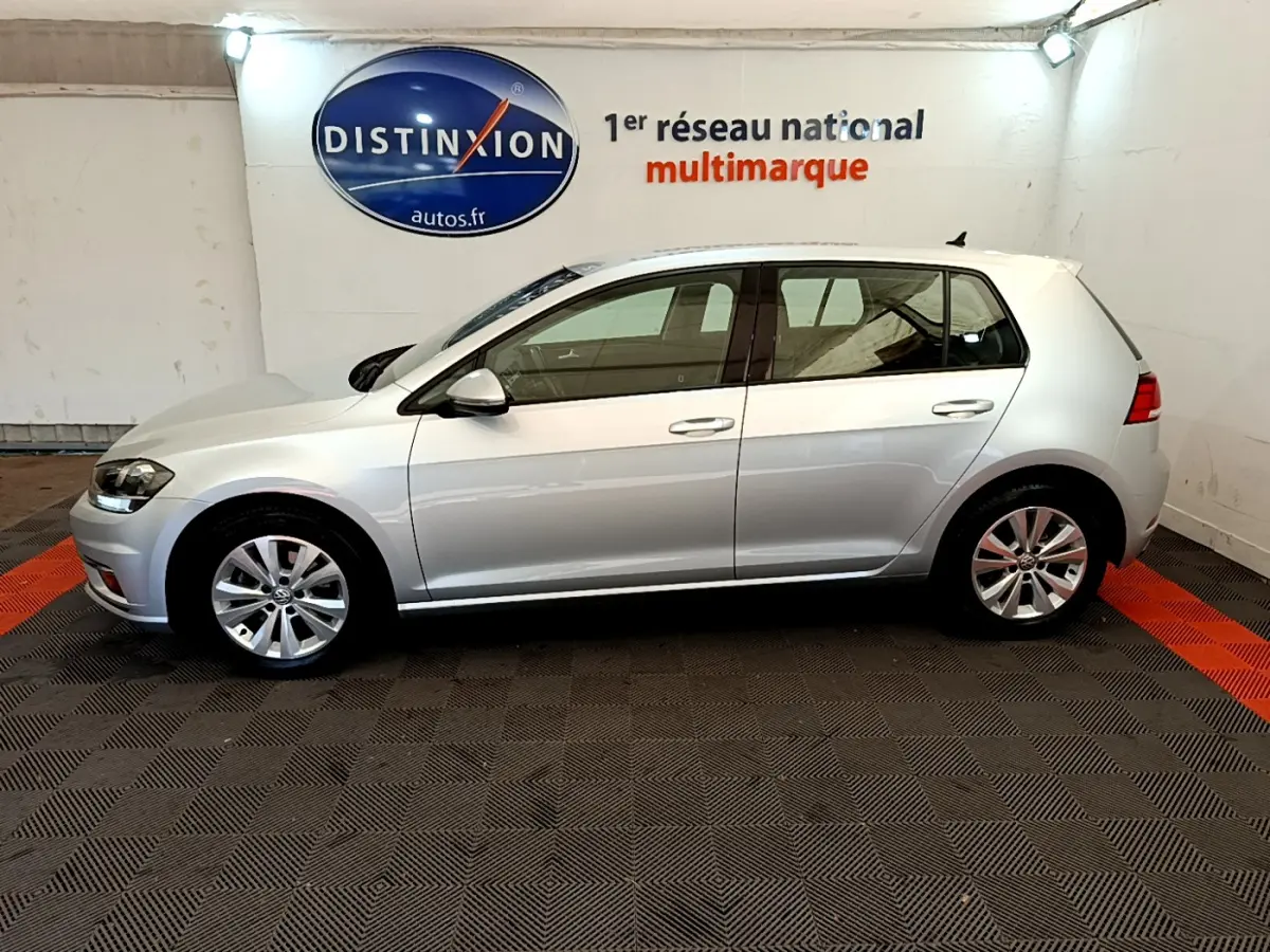 Profil droit d'une Volkswagen Golf gris clair 2019 garée en intérieur sur sol à damiers noir et orange.