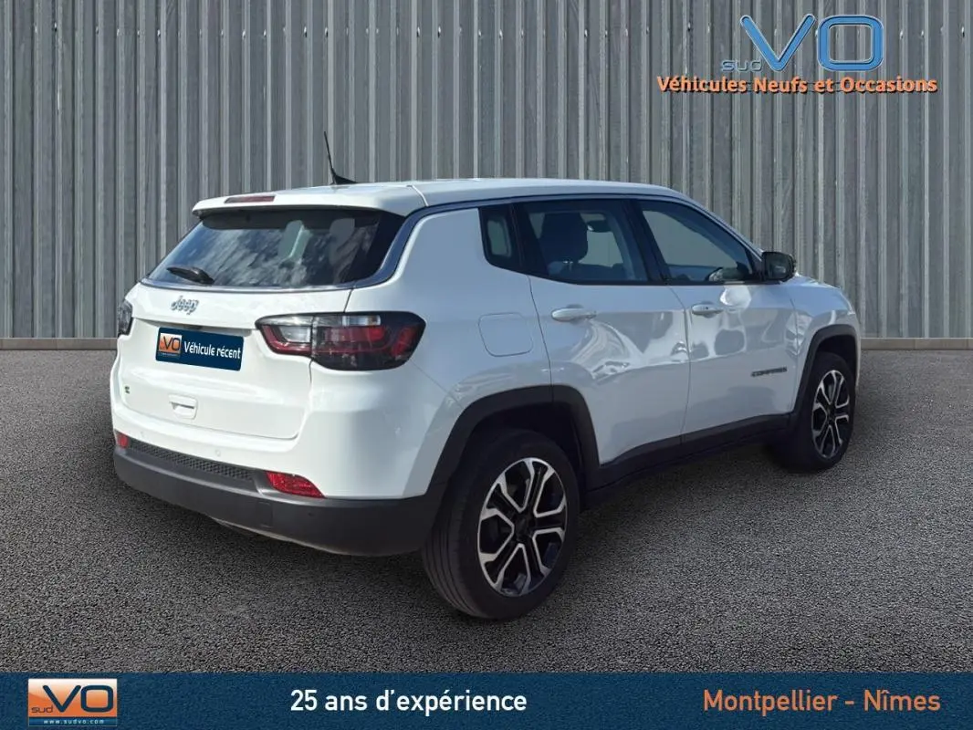 Jeep Compass blanc vue 3/4 arrière droit, jantes alliage 18 pouces, avec toit et vitres teintées, sur fond industriel.