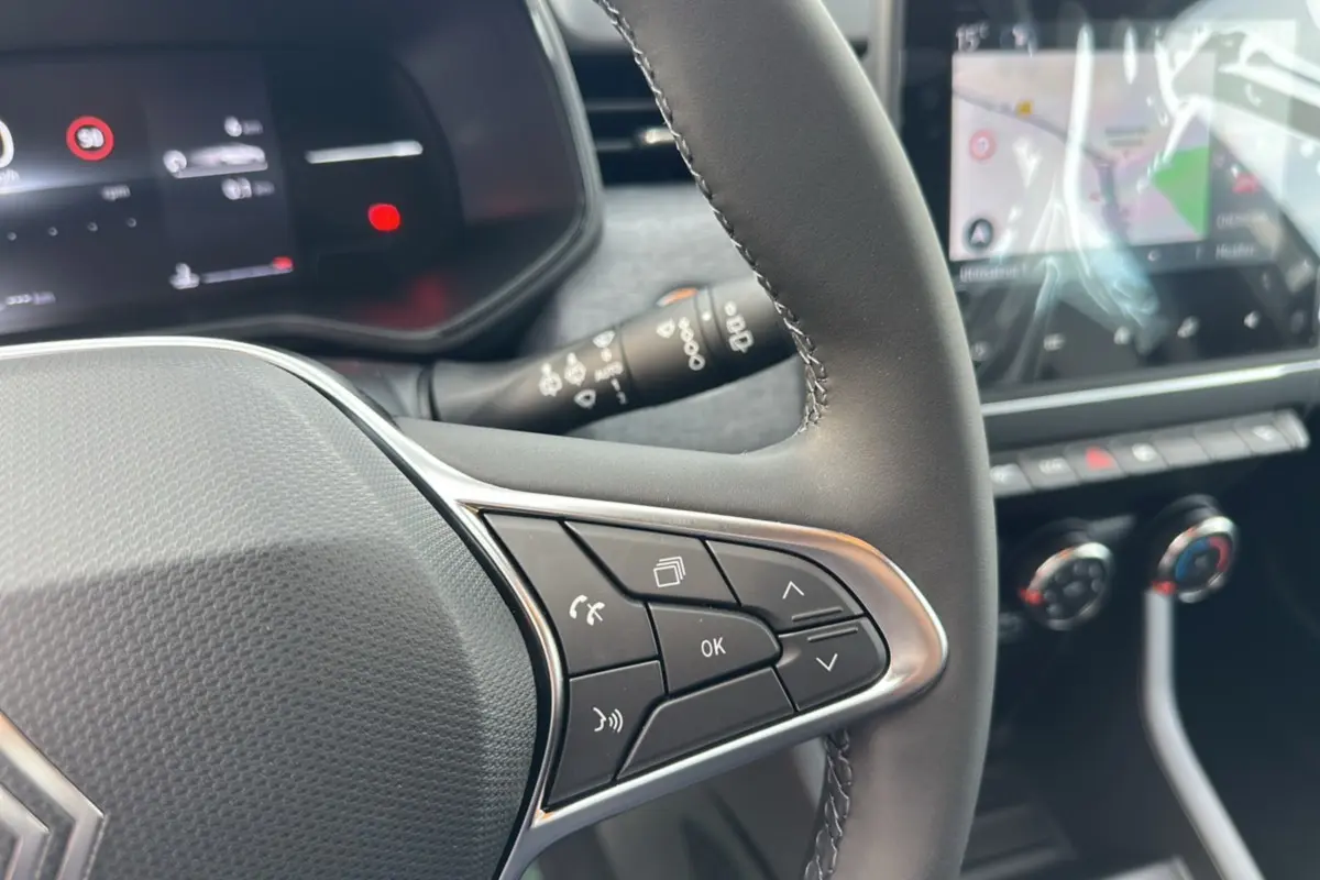 Gros plan sur le volant cuir multifonctions de la Renault Clio gris rafale, avec commandes intégrées et écran tactile en arrière-plan.