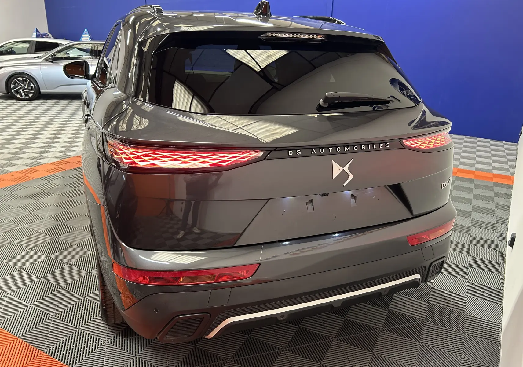 Vue arrière 3/4 d'un DS7 gris titane avec feux LED distinctifs et logo DS Automobiles sur fond intérieur showroom.