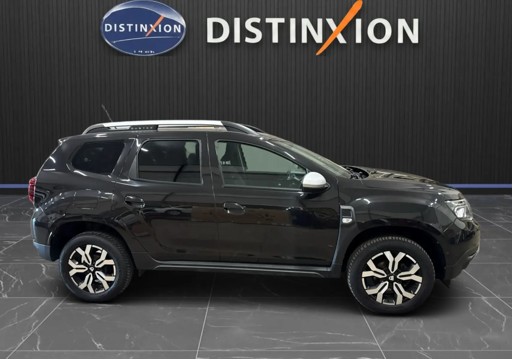 Vue de profil côté gauche d'un Dacia Duster noir 2022 avec jantes bi-ton et rétroviseurs blancs, sur fond intérieur showroom.