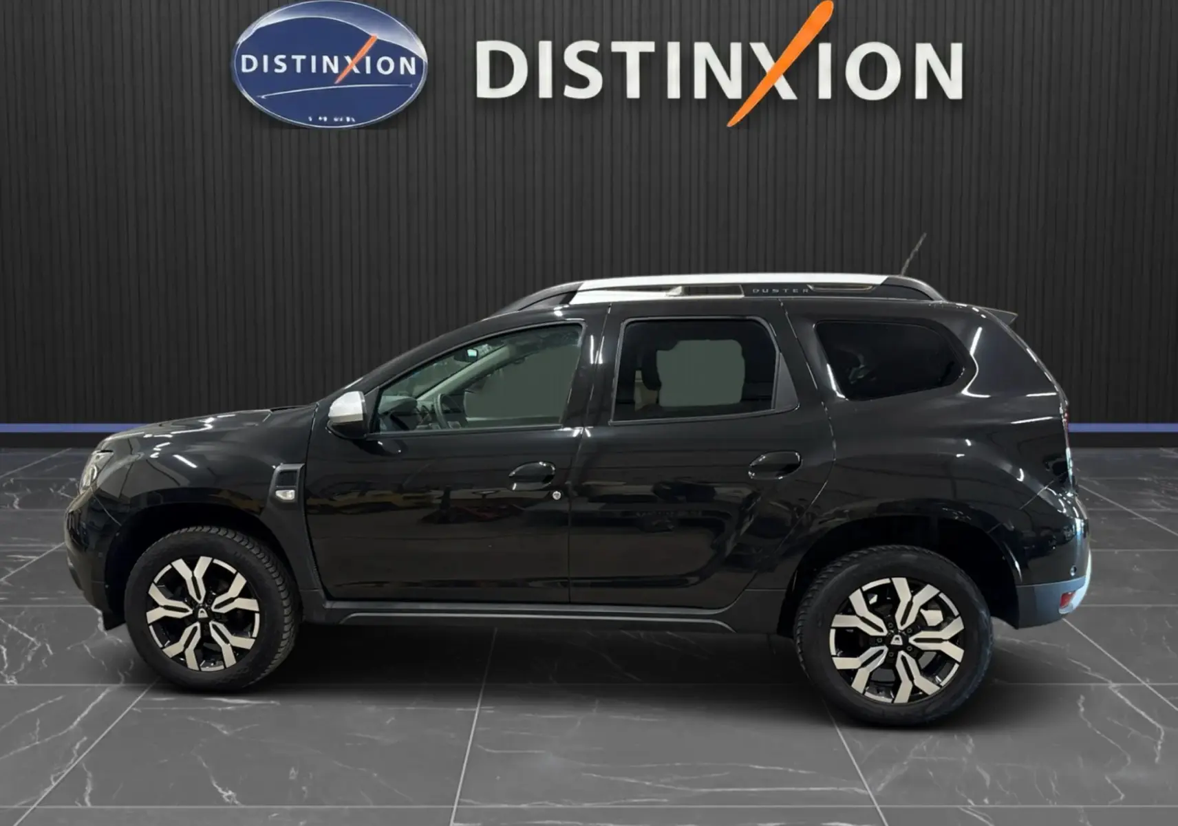 Profil côté gauche d'un Dacia Duster noir 2022 avec jantes bi-ton et barres de toit, sur fond intérieur showroom.
