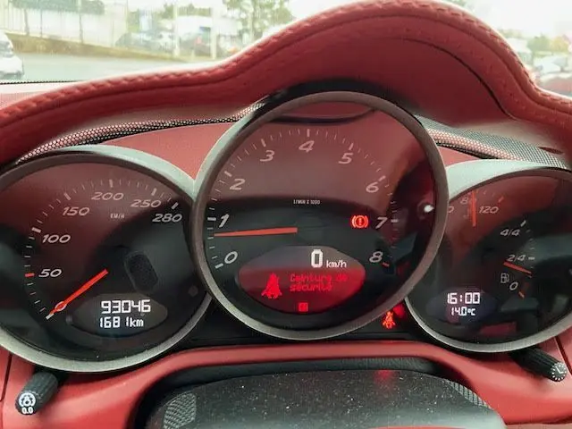 Vue rapprochée du tableau de bord rouge d'une Porsche Cayman noire 2008, affichant le compteur et alertes de sécurité.