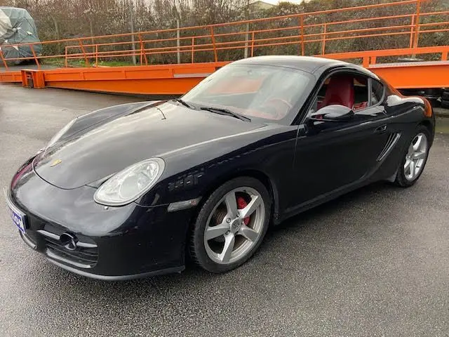 Porsche Cayman noir 2008 en 3/4 avant droit, jantes alu 5 branches et intérieur cuir rouge visible.