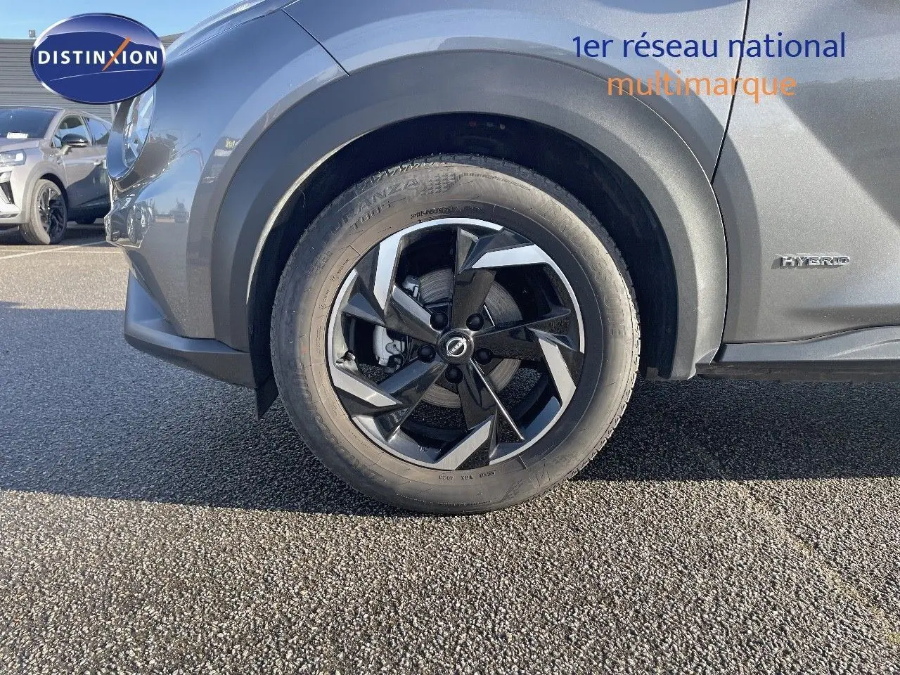 Gros plan sur la roue avant gauche du Nissan Juke gris squale métal 2024 avec jante alliage bicolore et badge Hybrid.