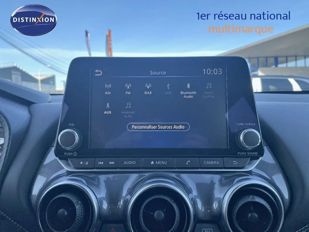 Écran tactile central du Nissan Juke gris squale métal 2024 affichant les sources audio, avec commandes et aérateurs visibles.