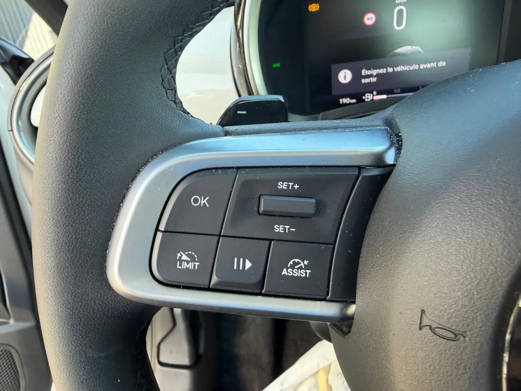 Gros plan sur les commandes du régulateur de vitesse au volant de la FIAT 600 blanc, tableau de bord numérique en arrière-plan.