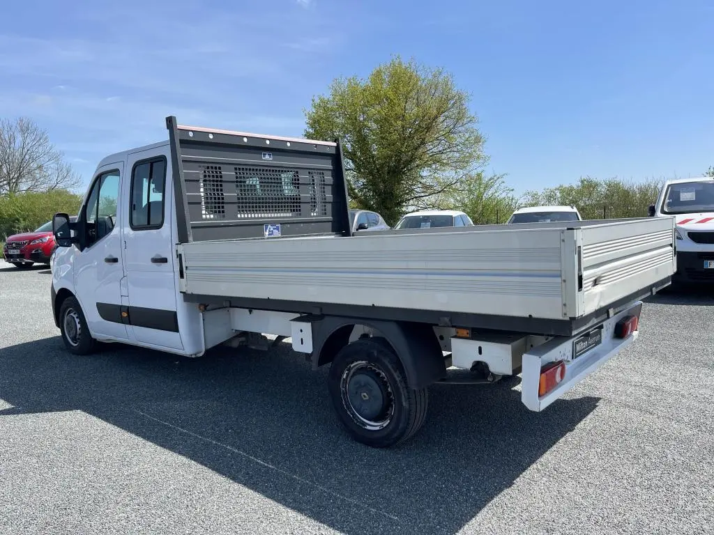 Renault Master blanc en 3/4 arrière droit, plateau ridelles aluminium et cabine double visible en extérieur sous ciel bleu.