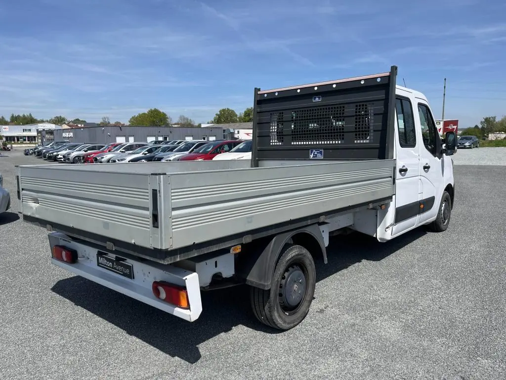 Renault Master blanc en vue 3/4 arrière droit, plateau ridelles aluminium et cabine double visible sur parking.