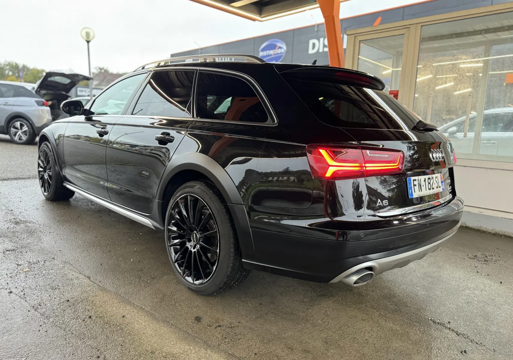 Audi A6 Avant Allroad noire vue 3/4 arrière droit, feux arrière allumés et jantes noires distinctives.
