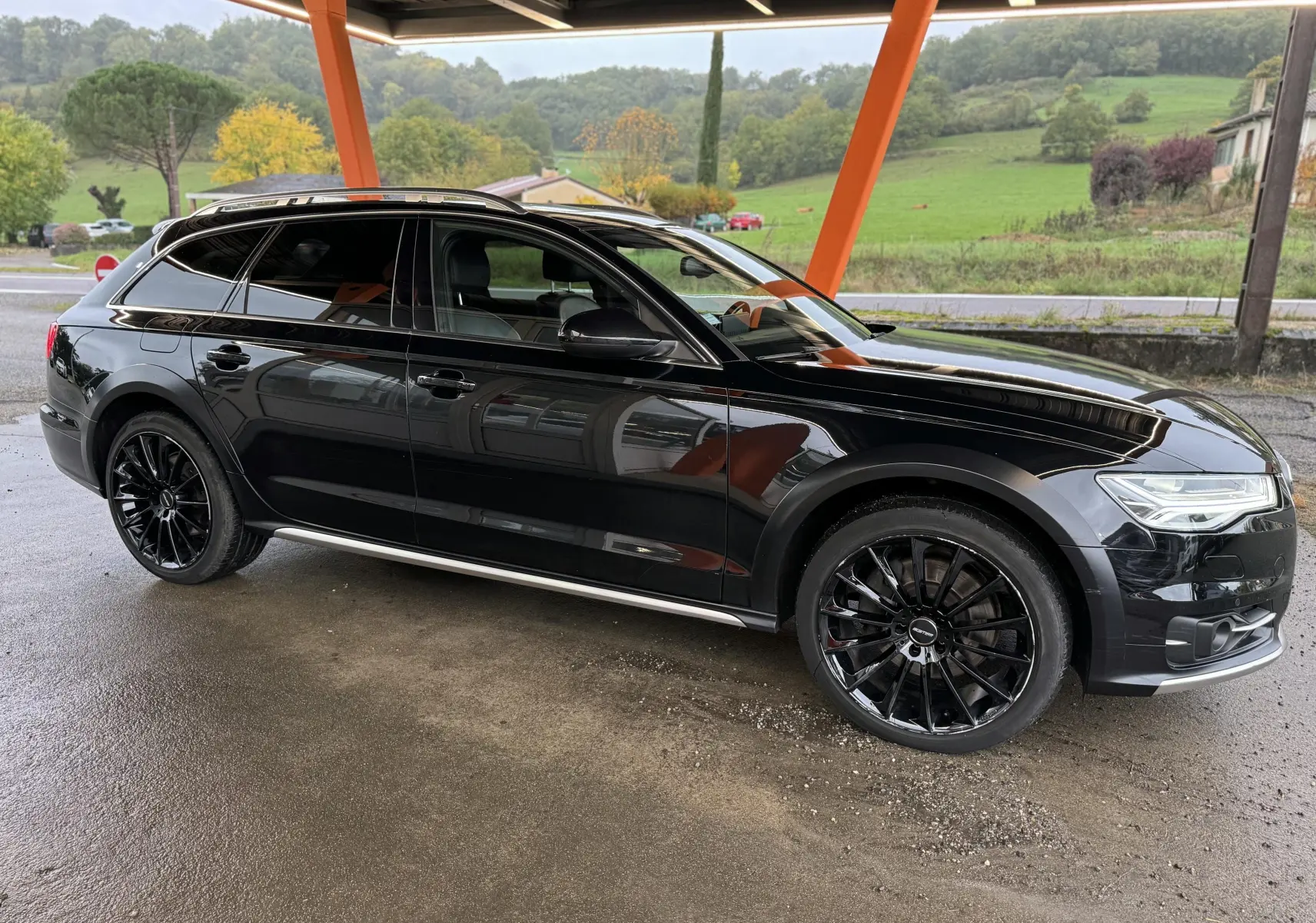 Audi A6 Avant Allroad noire vue de profil côté gauche, avec jantes noires et toit ouvrant sous abri.
