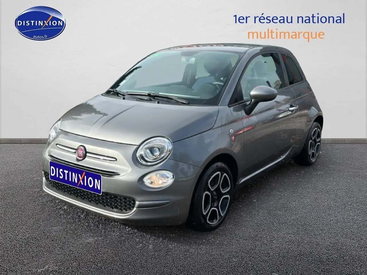 Fiat 500 gris vue 3/4 avant droit, avec jantes distinctives et phares ronds allumés.