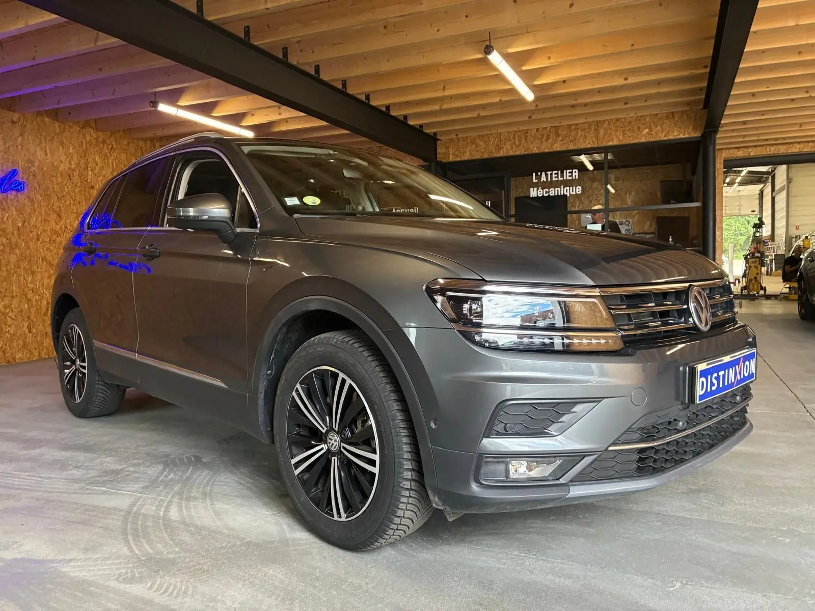 Volkswagen Tiguan gris 3/4 avant droit, avec jantes noires et détails chromés, stationné en intérieur d'atelier.