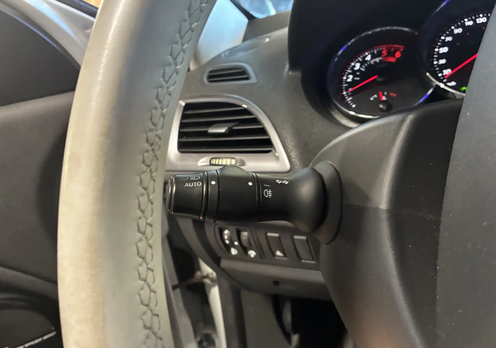 Gros plan sur le volant cuir et la commande des phares automatiques à gauche du tableau de bord d'une Renault Mégane Coupé blanche.