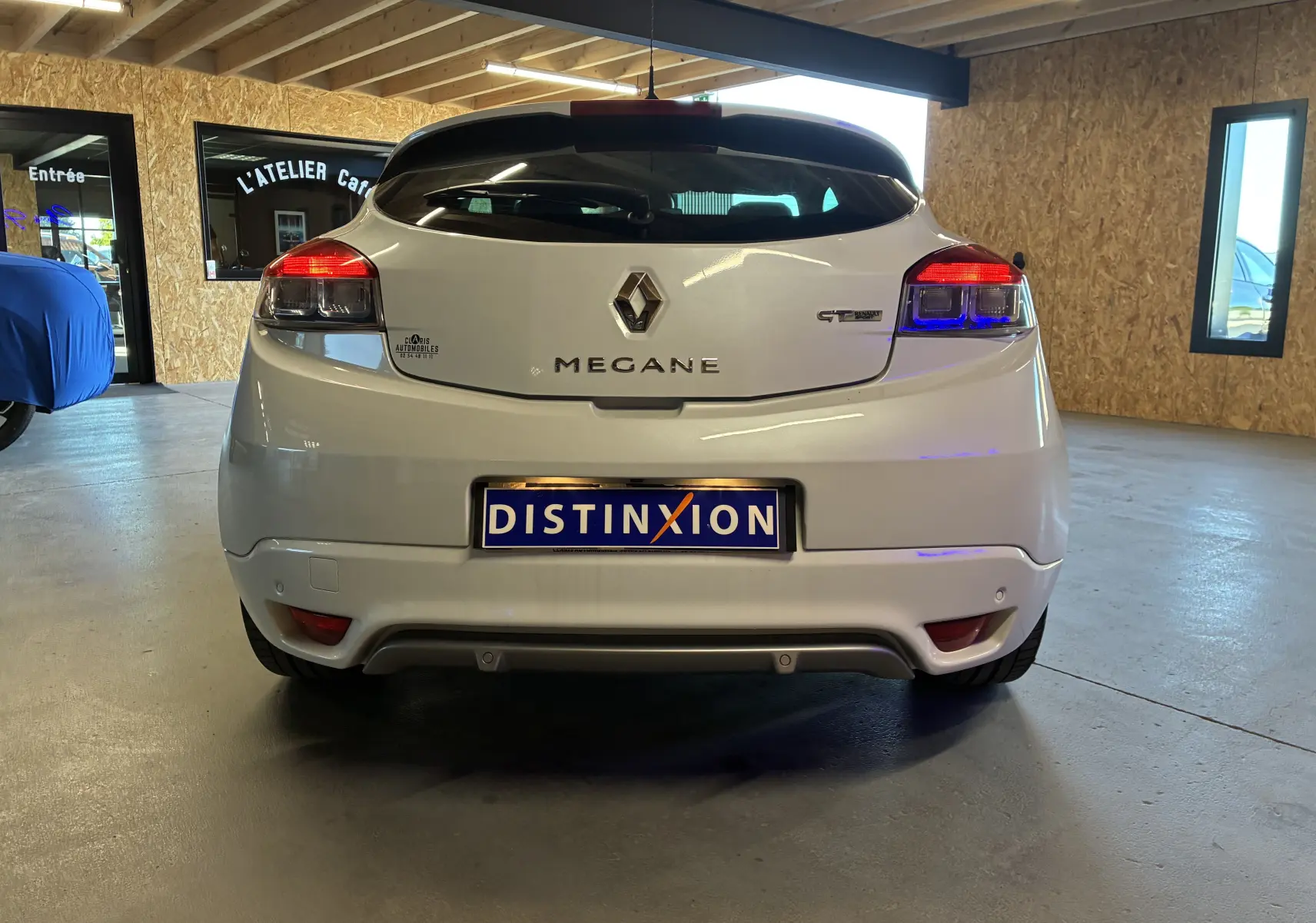 Vue arrière d'une Renault Mégane Coupé 2.0 DCI 165 GT blanche, avec feux arrière allumés, en intérieur.