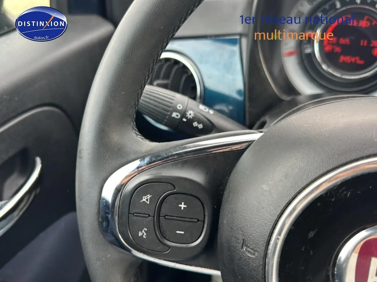 Gros plan sur le volant multifonction noir de la Fiat 500 bleu foncé, avec commandes audio et tableau de bord partiel visible.