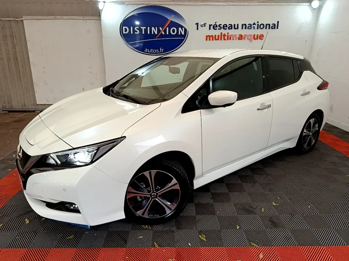 Vue 3/4 avant gauche d'une Nissan Leaf électrique blanche avec jantes alliage et phares LED allumés en showroom.