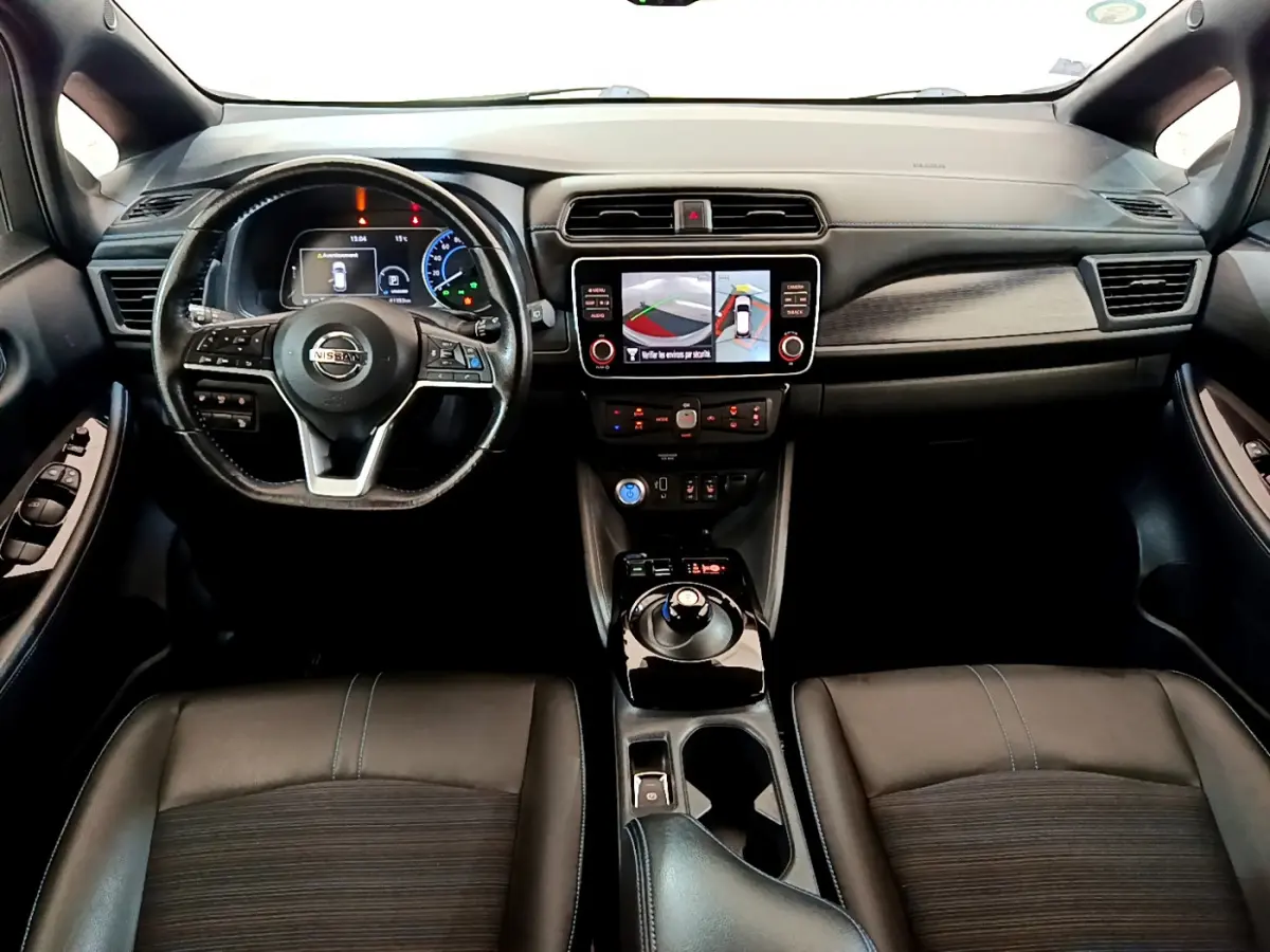 Vue intérieure avant du Nissan Leaf blanc 2020, volant cuir, écran tactile central affichant la caméra 360° d'aide au stationnement.