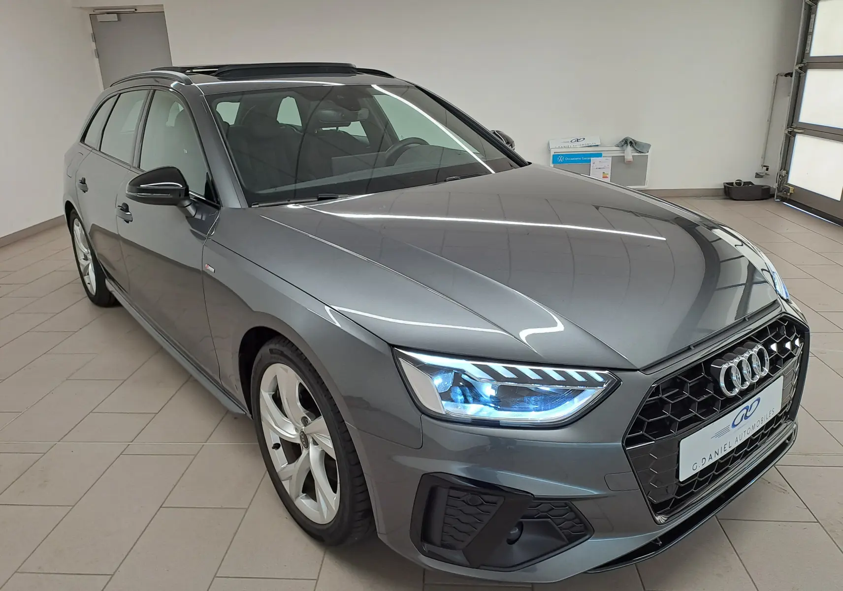 Audi A4 Avant gris Daytona vue 3/4 avant droit en intérieur avec phares LED allumés et toit noir panoramique