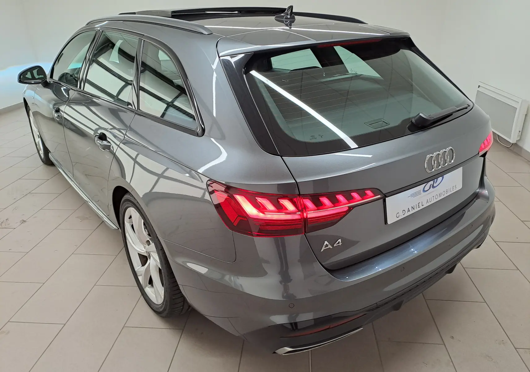 Audi A4 Avant gris Daytona vue 3/4 arrière droit, avec toit panoramique et feux arrière LED allumés.