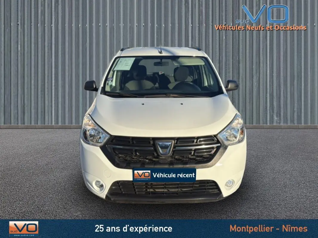 Vue frontale d'un Dacia Lodgy blanc avec calandre noire et barres de toit, stationné devant un mur métallique.