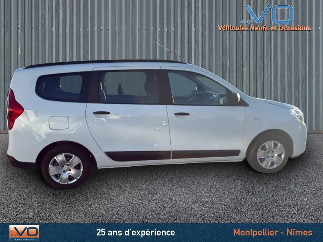 Profil droit du Dacia Lodgy blanc 2018 avec barres de toit, vitres teintées et jantes acier en arrière-plan urbain.