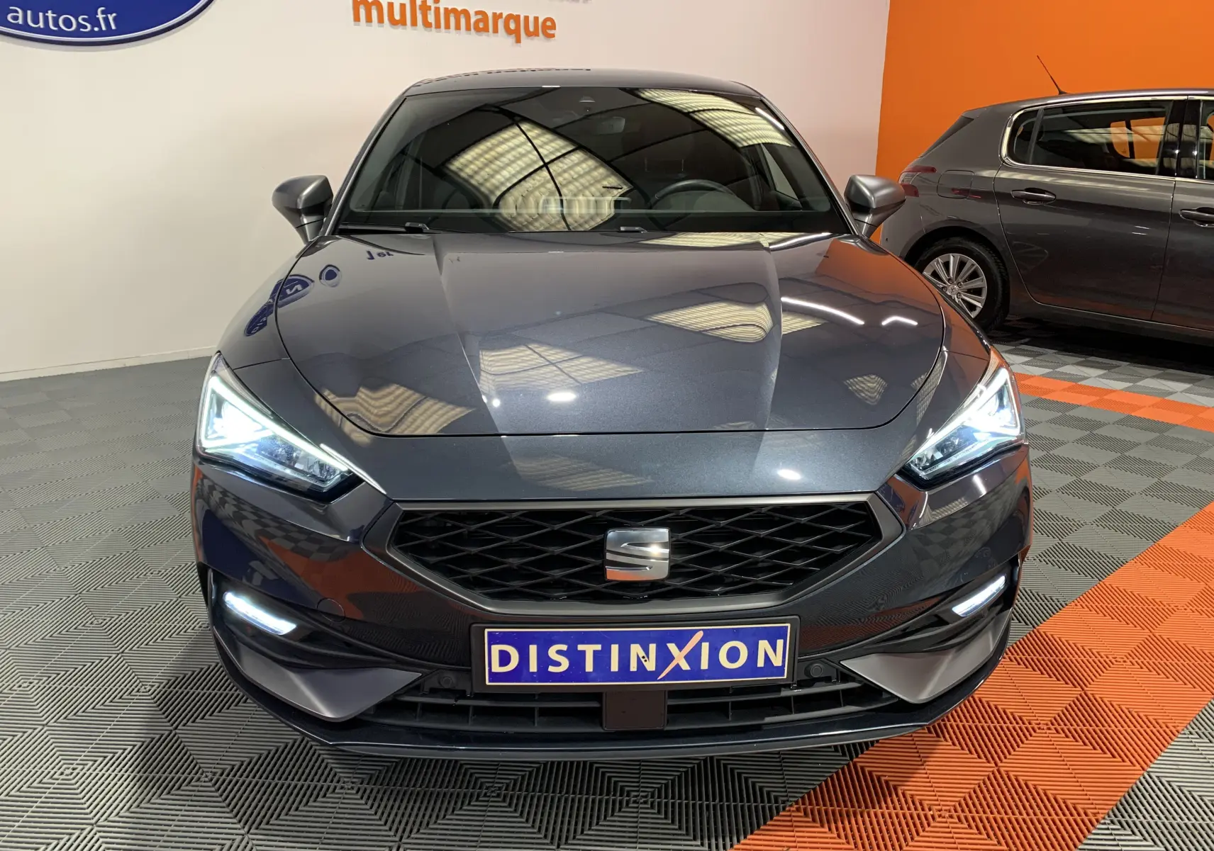 Vue frontale d'une SEAT LEON E-HYBRID 204 FR gris métallisé avec phares LED allumés en intérieur showroom.