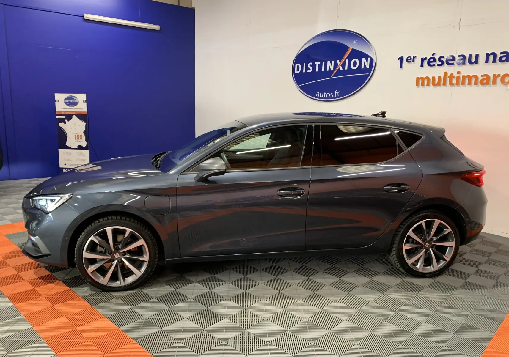 Profil côté gauche d'une SEAT LEON E-HYBRID 204 FR gris métallisé, avec jantes alliage et vitres teintées, en intérieur showroom.