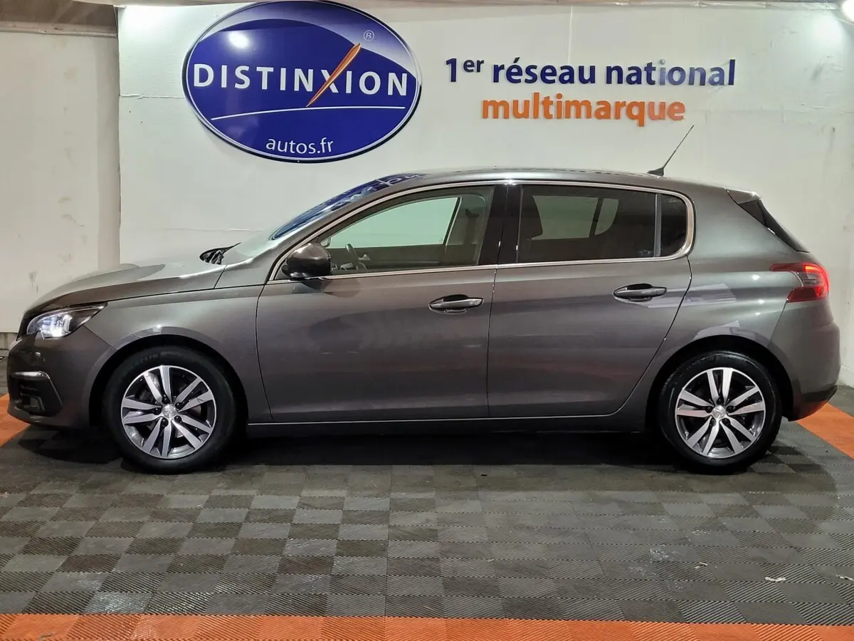 Profil côté gauche d'une Peugeot 308 gris foncé 2020 garée en intérieur sur sol damier, avec jantes alliage visibles.