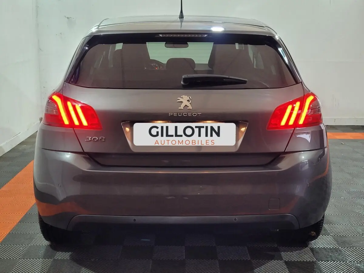Vue arrière d'une Peugeot 308 gris foncé avec feux arrière allumés et logo visible sur un sol damier noir et orange.