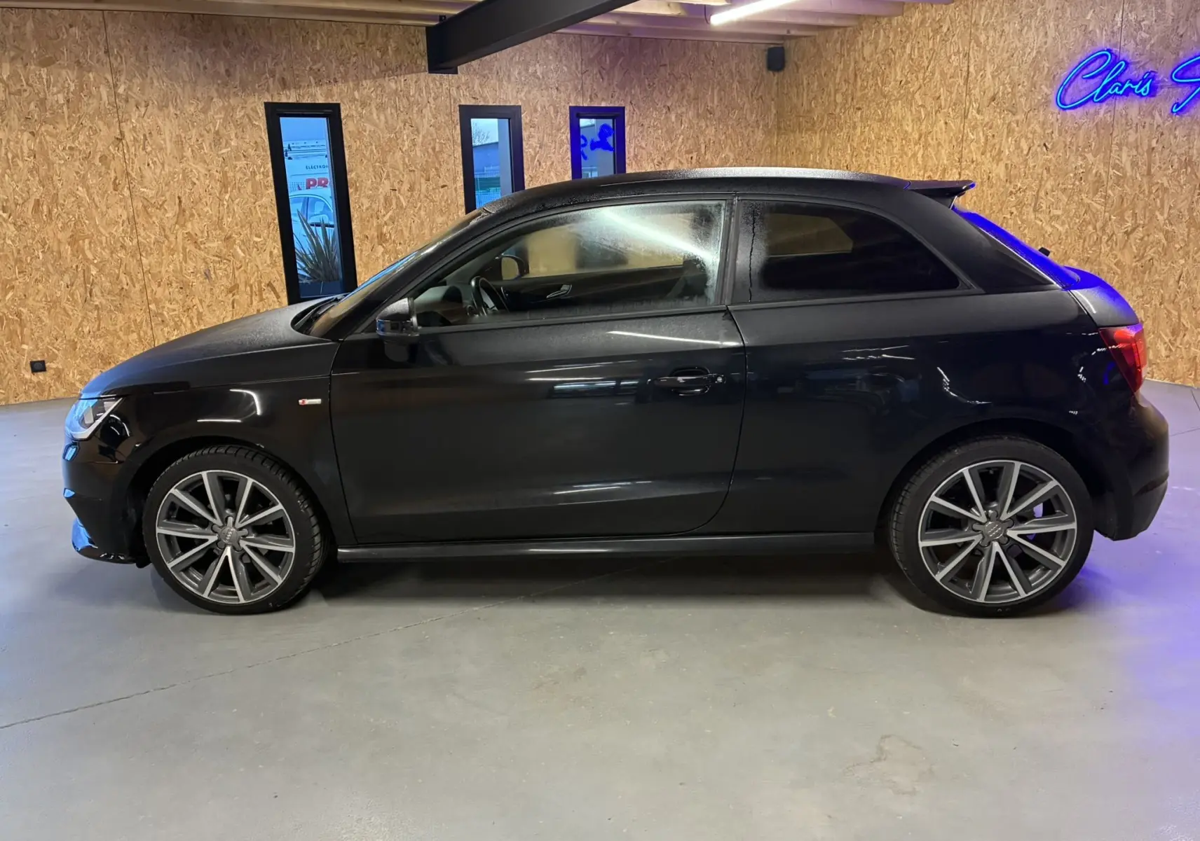 Profil côté gauche d'une Audi A1 noire 3 portes, version S-Line avec jantes alliage 17 pouces dans un intérieur bois.