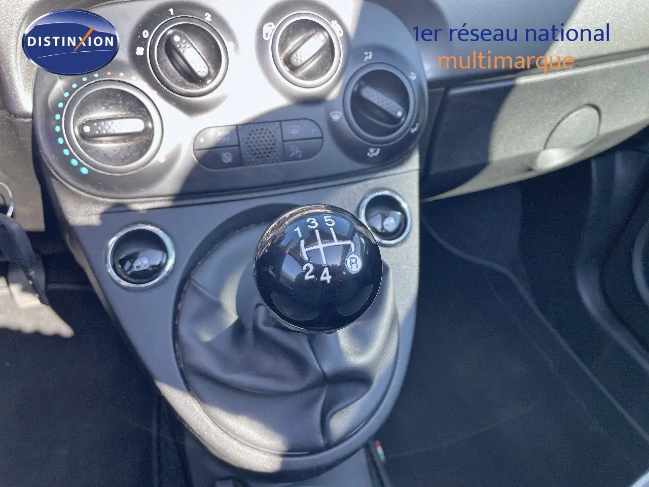 Gros plan sur le levier de vitesses manuel noir dans l'habitacle d'une Fiat 500C grise, avec commandes de climatisation visibles.