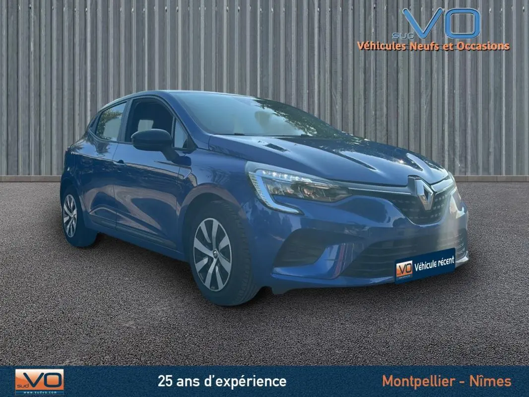 Renault Clio V TCe 90 Equilibre bleu foncé vue 3/4 avant droit sur sol bitumé devant un mur métallique.