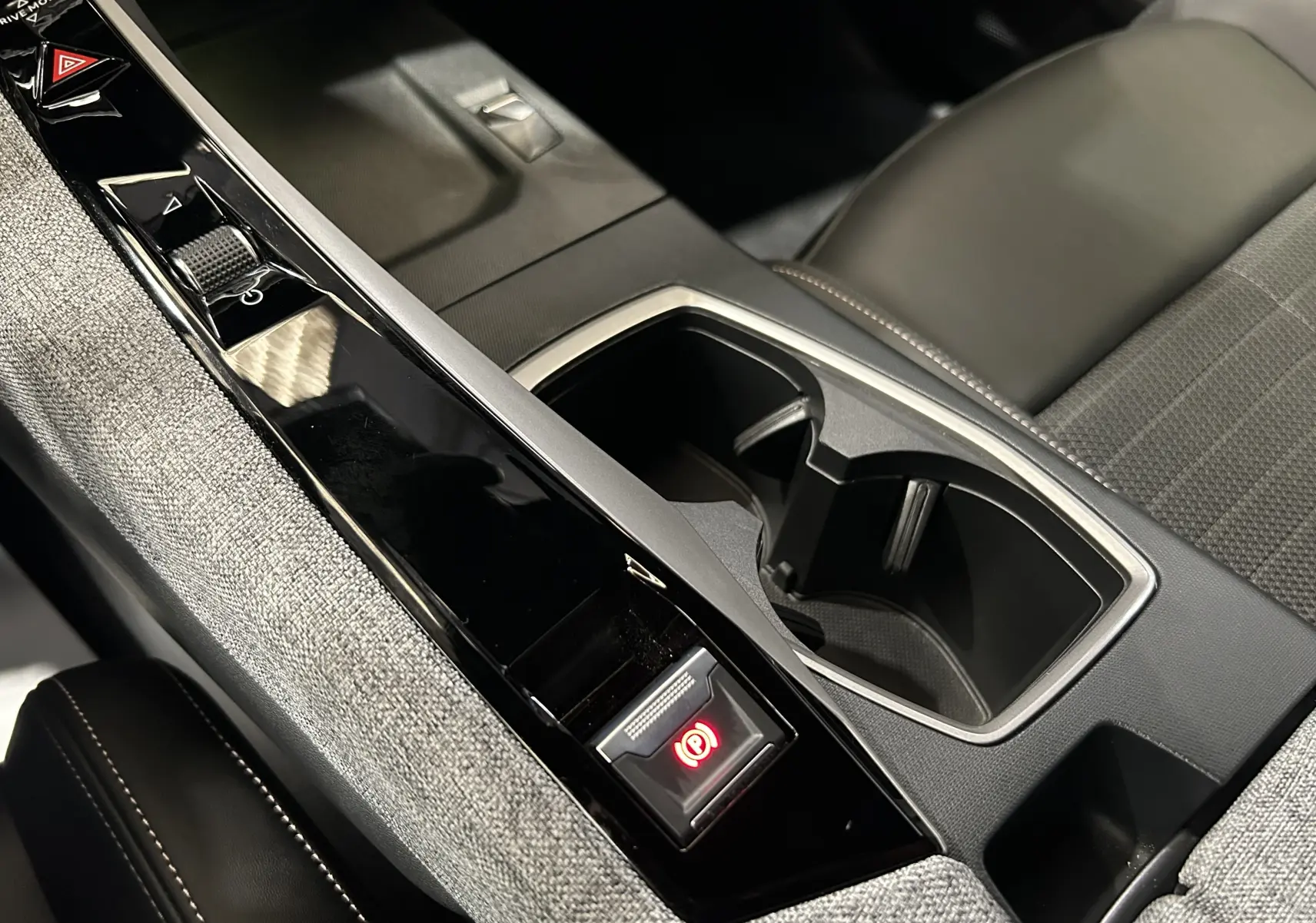 Vue rapprochée de la console centrale du Peugeot 3008 Hybrid 2024, avec tissu gris et bouton frein à main électronique rouge.