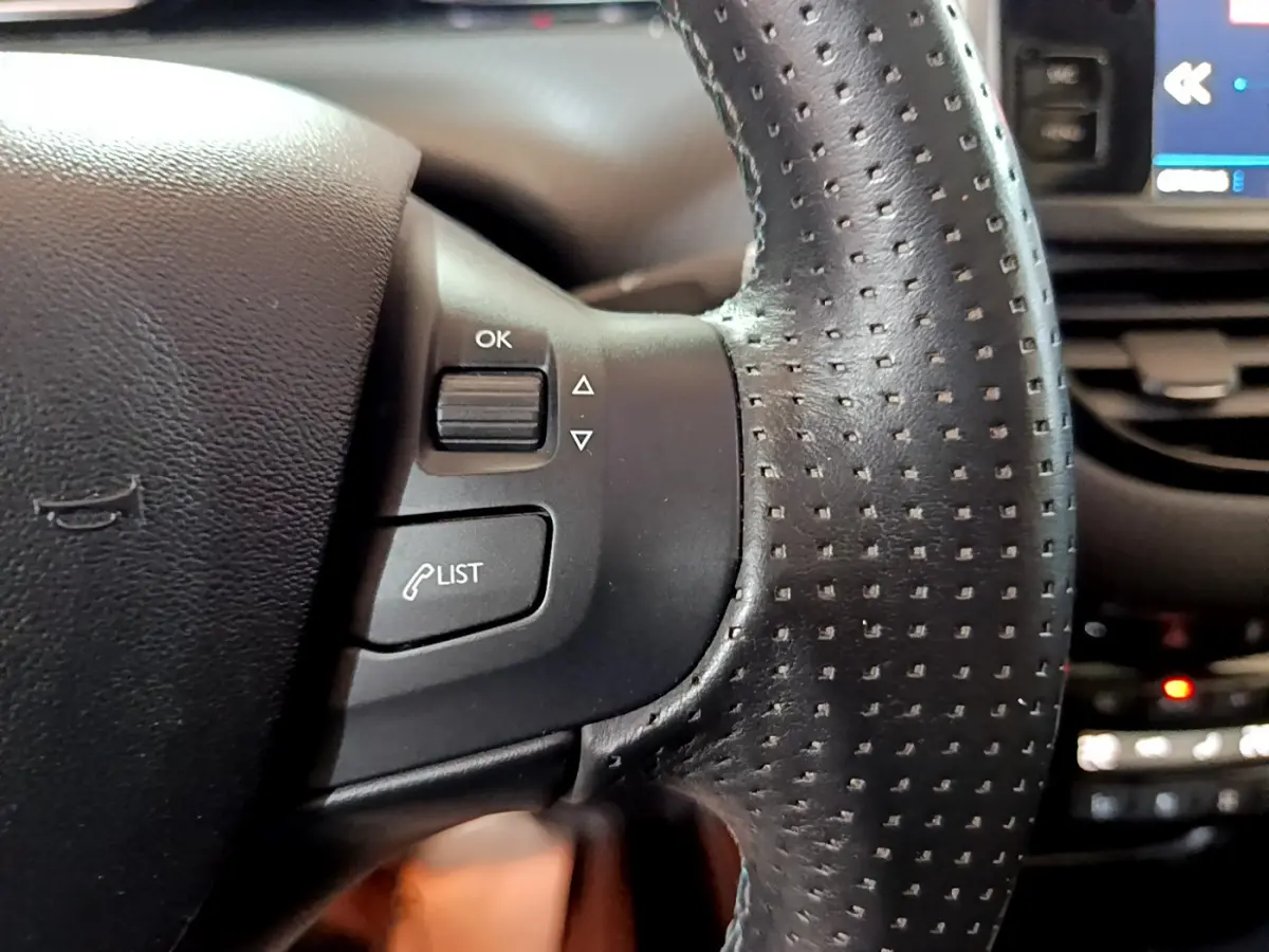 Gros plan sur les commandes gauche du volant en cuir perforé du Peugeot 2008 gris foncé, avec écran tactile en arrière-plan.