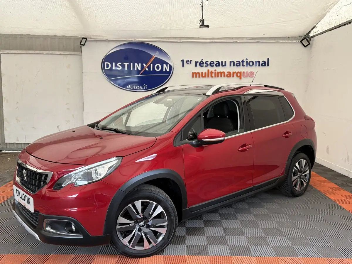 Peugeot 2008 rouge vue 3/4 avant droit, avec jantes alliage et barres de toit chromées en intérieur showroom.